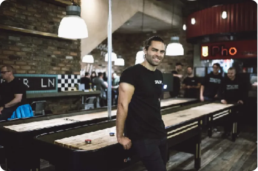 A member of the SHUFL team standing beside a shuffleboard table in a busy venue