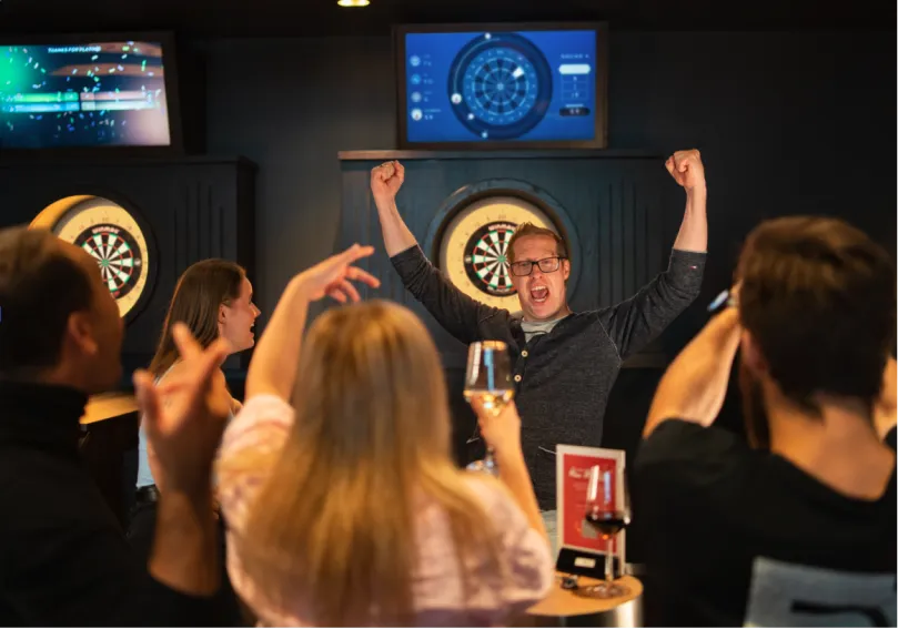A group cheering with their arms in the air in front of an interactive darts oche
