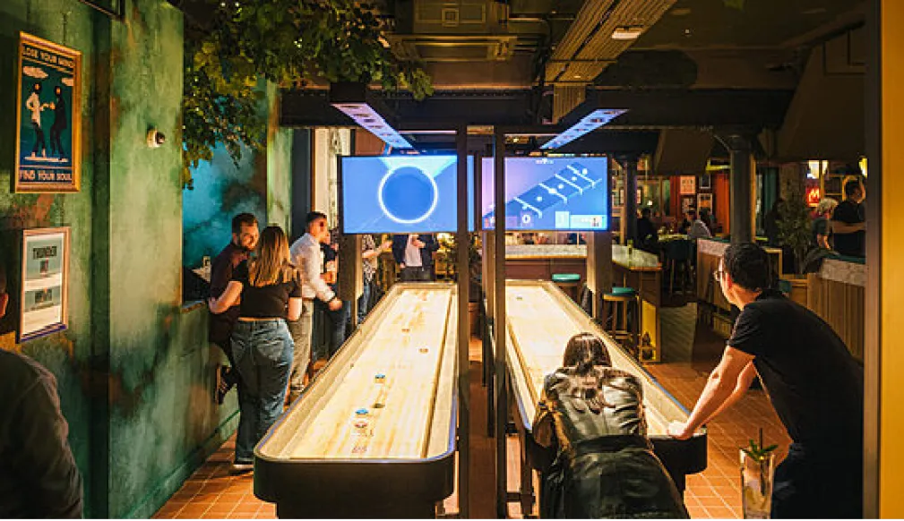 Guests playing shuffleboard in a plant-filled bar with screens above the table